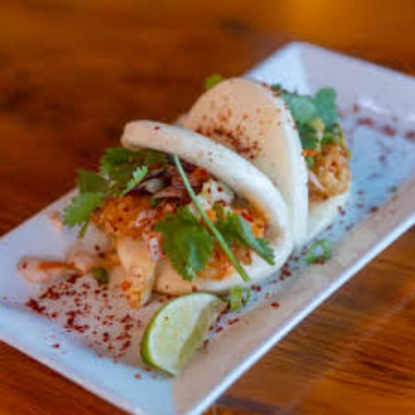 Two bao buns filled with fried chicken, slaw, cilantro, and chili flakes, served with a lime wedge.