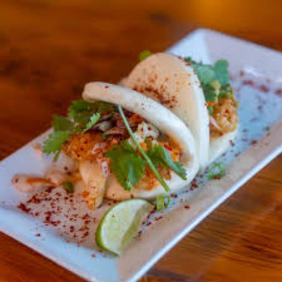 Two bao buns filled with fried chicken, slaw, cilantro, and chili flakes, served with a lime wedge.