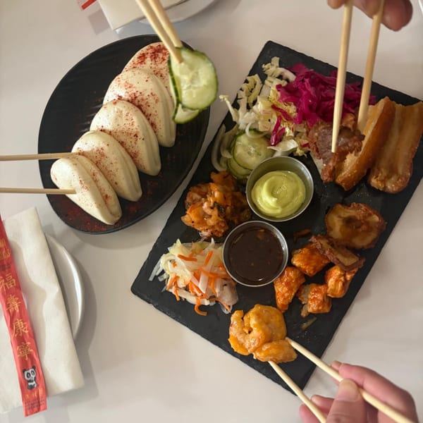 A black platter with various Asian appetizers, including bao buns, pork belly, shrimp, and pickled vegetables.