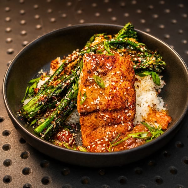 Seared salmon with sesame seeds, roasted asparagus, white rice, and sauce in a dark bowl.