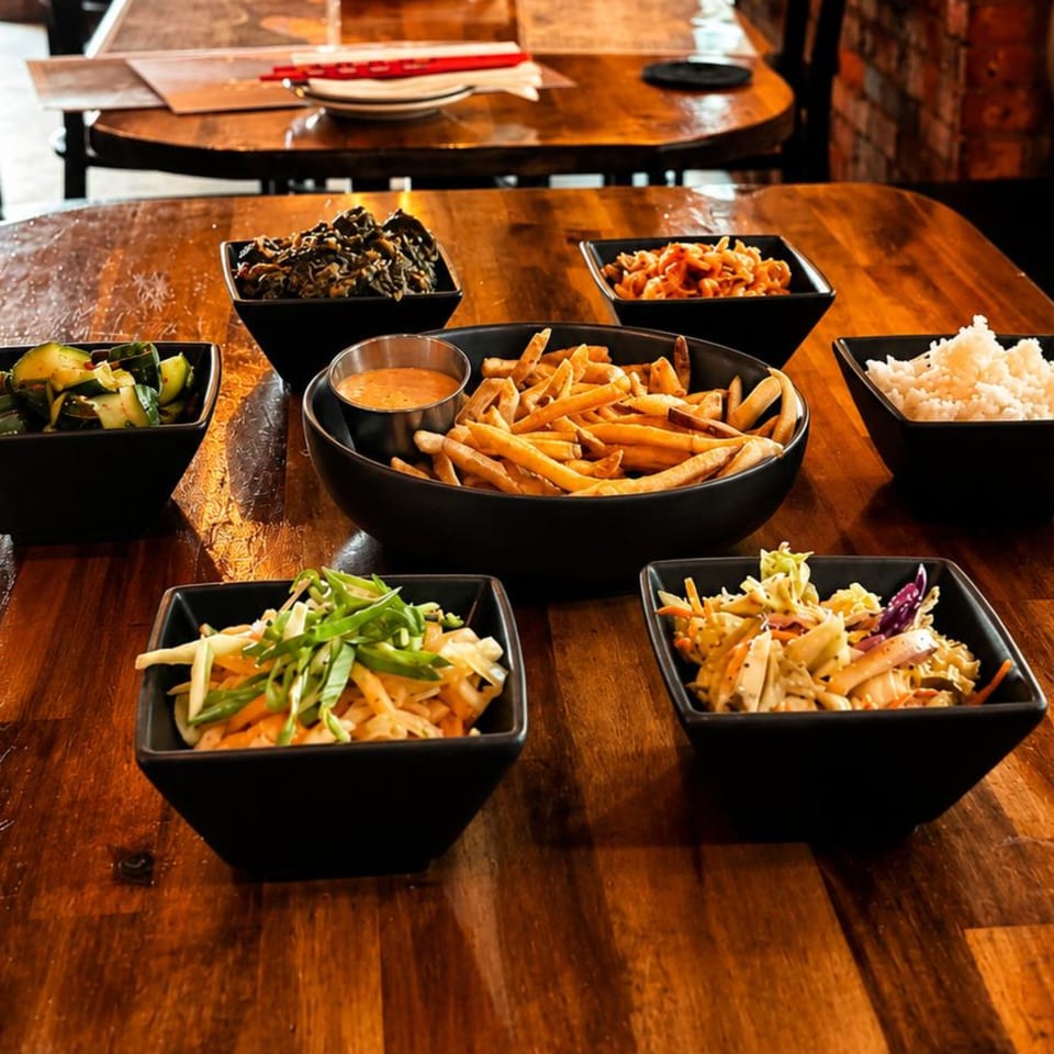 An assortment of Asian side dishes, including fries, rice, and various salads, on a wooden table.